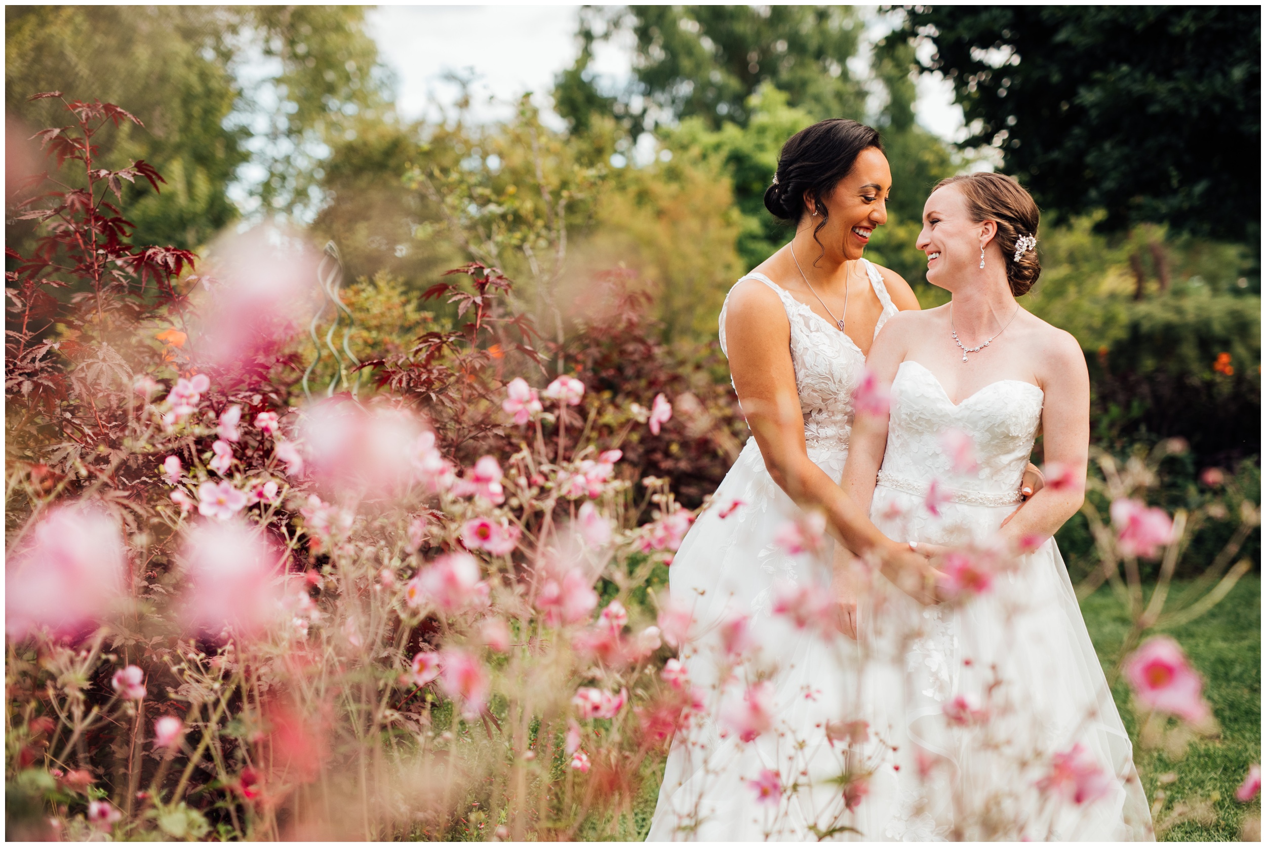 Two brides embracing among pink flowers at New England Botanic Garden at Tower Hill during their same sex wedding